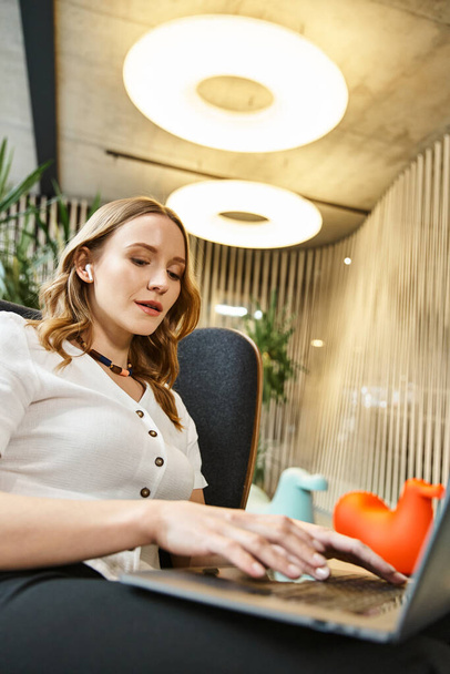 Eine Frau, die mitten in der Arbeit auf einem Stuhl sitzt und einen Laptop in einem modernen Coworking Space benutzt. - Foto, Bild