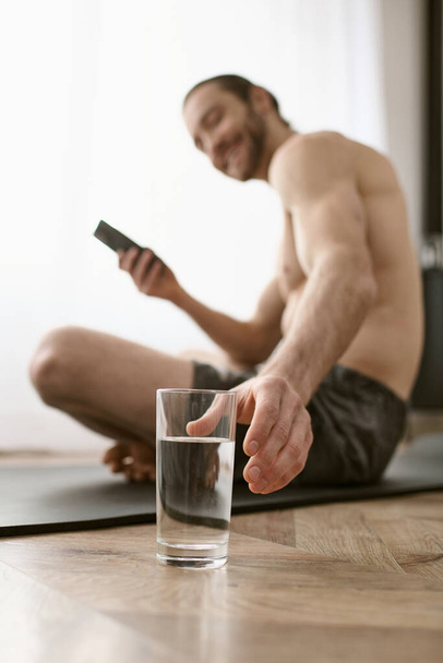 Ένας άντρας κάθεται στο πάτωμα, κρατώντας ένα ποτήρι νερό και ένα κινητό τηλέφωνο.. - Φωτογραφία, εικόνα