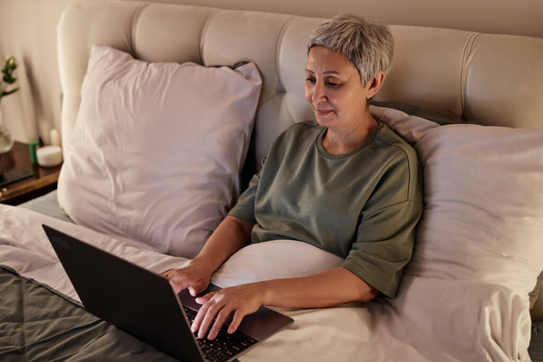 Hoge hoek portret van Aziatische senior vrouw met behulp van laptop terwijl liggend in comfortabel bed thuis - Foto, afbeelding