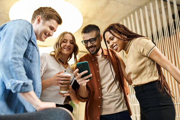 Un groupe diversifié de collègues dans un espace de coworking moderne sont réunis autour d'un téléphone portable, absorbé par ce qu'ils voient. - Photo, image