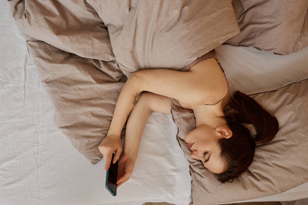 Retrato de vista superior mínimo da jovem deitada na cama confortável e usando o smartphone rolando mídias sociais, espaço de cópia - Foto, Imagem