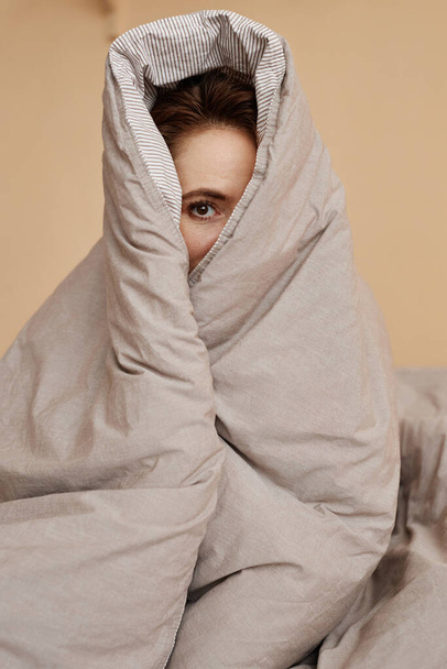 Portrait of cute young woman wrapped in cozy blanket peeking at camera with one eye - Photo, Image