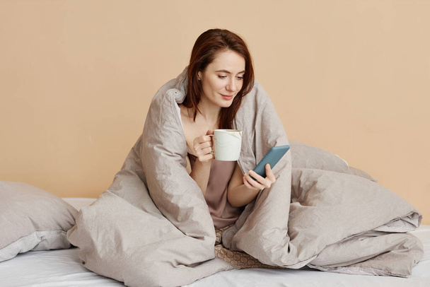Retrato de sorrindo jovem coberta de cobertor acolhedor desfrutando de fim de semana relaxante em casa e segurando o telefone, espaço de cópia - Foto, Imagem