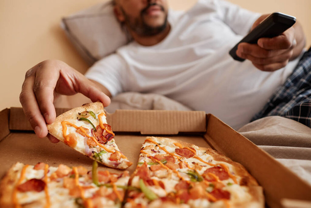 Nahaufnahme eines erwachsenen Mannes, der ein Stück Pizza nimmt, während er ein gemütliches Wochenende zu Hause genießt und fernsieht, Kopierraum - Foto, Bild