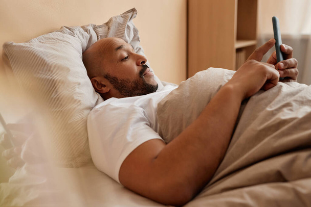 Zijaanzicht portret van zwarte volwassen man met behulp van smartphone in bed thuis in luie ochtend - Foto, afbeelding