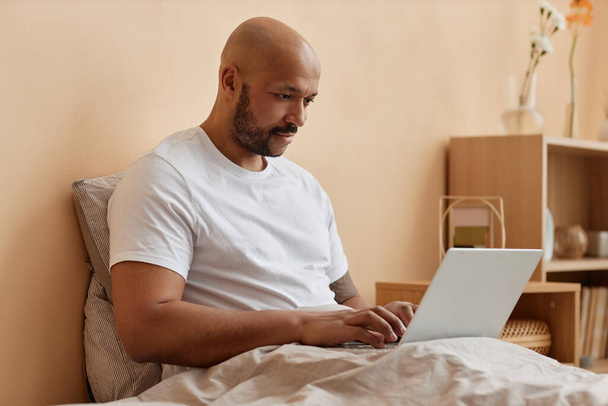 Retrato de la vista lateral del hombre negro calvo usando el ordenador portátil en la cama en el hogar acogedor, espacio de copia - Foto, imagen