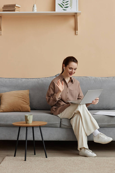 Retrato vertical de larga duración de una joven mujer de negocios elegante disfrutando del trabajo desde casa y sentada en un cómodo sofá saludando a una reunión en línea - Foto, imagen