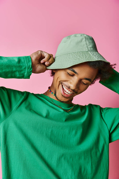 A cheerful young African American man with curly hair wearing a green shirt and green hat on a pink background. - Photo, Image