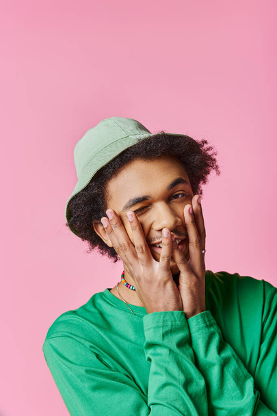 Un jeune homme afro-américain joyeux et frisé porte une chemise verte et un chapeau sur un fond rose. - Photo, image