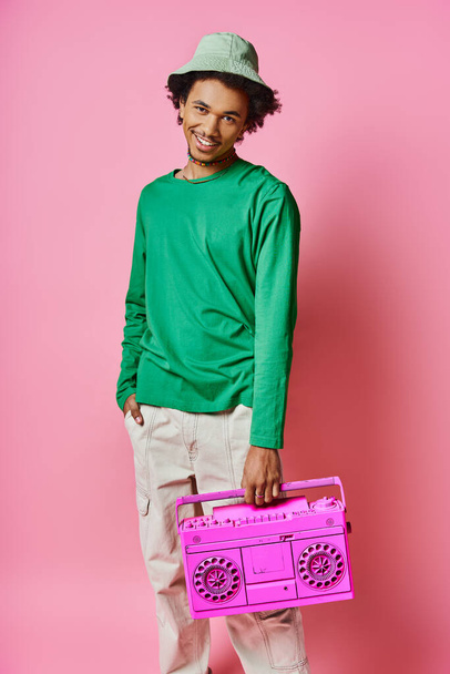 Uomo afroamericano riccio con una camicia verde che tiene gioiosamente una radio rosa su uno sfondo rosa. - Foto, immagini