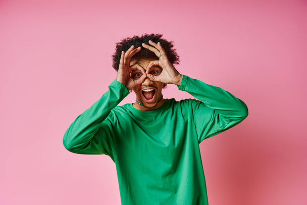 Un hombre afroamericano alegre con el pelo rizado en una camisa verde, sosteniendo alegremente sus manos hacia arriba a su cara sobre un fondo rosado. - Foto, Imagen