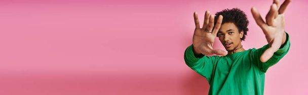 Cheerful curly African American man in casual green shirt with hands raised, expressing positivity on a pink background. - Photo, Image