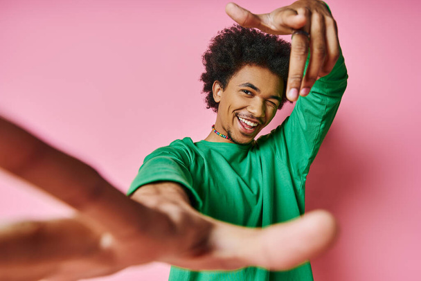 Un homme afro-américain joyeux, cheveux bouclés dans une chemise verte, danse énergiquement sur un fond rose vif. - Photo, image