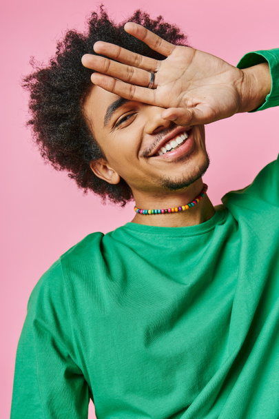 Een vrolijke jonge Afro-Amerikaanse man in een groen shirt, die zijn hand tegen zijn gezicht houdt en een emotioneel gebaar toont.. - Foto, afbeelding