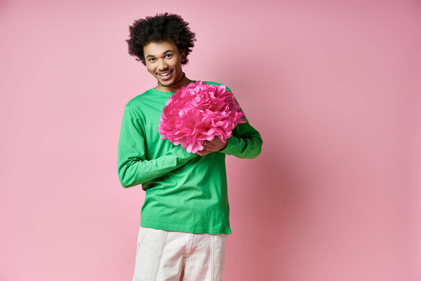 Homem afro-americano alegre em camisa verde segurando uma flor rosa, expressando emoções em um fundo rosa. - Foto, Imagem