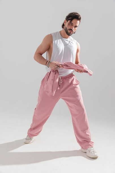 ハッケルを持っているピンクのスポーツ服を着ているセクシーな筋肉男性の完全なボディ写真,腕を示し,灰色の背景の前に歩く - 写真・画像