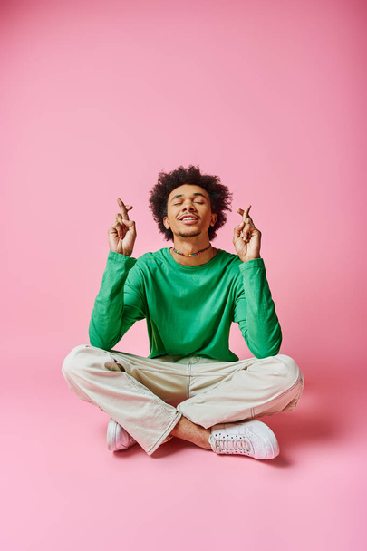 A cheerful young African American man in a green shirt strikes a yoga pose with focus and grace on a pink background. - Photo, Image
