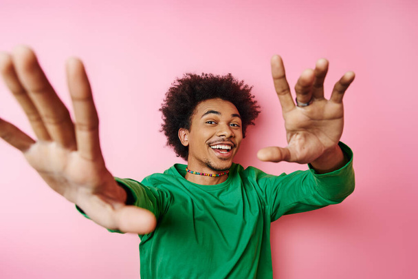 Young, curly-haired man in casual attire, raising his hands in excitement on a pink background. - Photo, Image