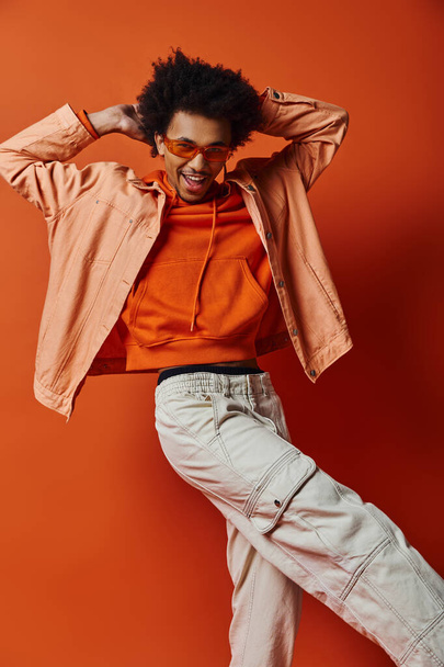 Stylový mladý Afroameričan v oranžové košili a bílých kalhotách, nosí sluneční brýle, vyzařující sebevědomí na oranžovém pozadí. - Fotografie, Obrázek