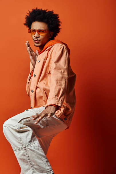 Curly African American man standing gracefully on one leg, exuding confidence and style, wearing trendy attire, on orange background. - Photo, Image