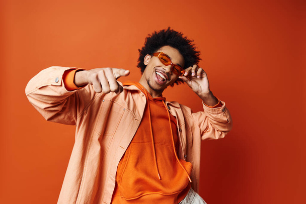 Un joven y elegante hombre afroamericano con el pelo rizado con una camisa naranja y gafas de sol, posando enérgicamente sobre un fondo naranja. - Foto, Imagen