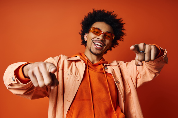 Stilvoller afroamerikanischer Mann in orangefarbenem Hemd und Sonnenbrille, der auf leuchtend orangefarbenem Hintergrund in Richtung Kamera zeigt. - Foto, Bild