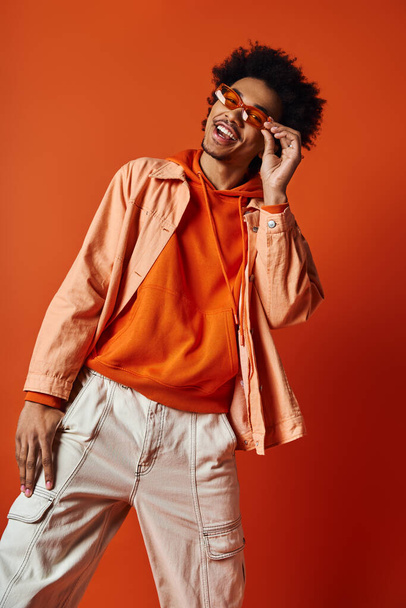 Afro-Amerikaanse man met krullend afro haar en een zonnebril die expressief poseert in een oranje shirt. - Foto, afbeelding