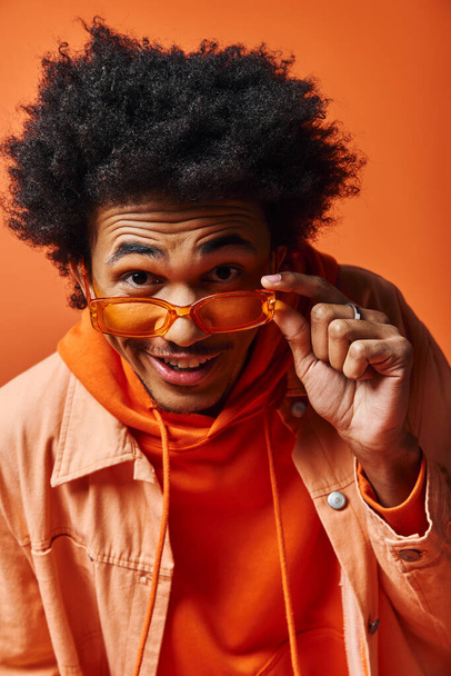Hombre afroamericano de moda con pelo afro rizado y gafas de sol posando expresivamente en una camisa naranja. - Foto, imagen
