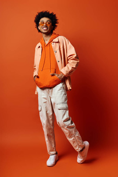Giovane uomo afroamericano in abiti alla moda e occhiali da sole, esprimendo emozioni su sfondo arancione. - Foto, immagini