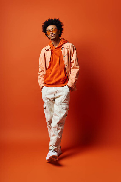 Stilvoller junger afroamerikanischer Mann in trendiger Kleidung und Sonnenbrille, der Emotionen vor auffallend orangefarbener Kulisse ausstrahlt. - Foto, Bild