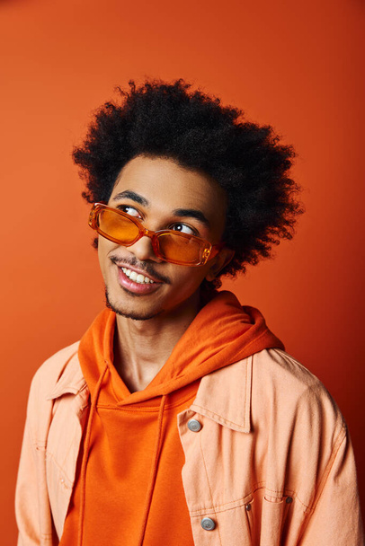 Egy divatos afro-amerikai férfi divatos kabátot és napszemüveget visel, magabiztosságot áraszt a narancssárga háttér ellenére.. - Fotó, kép