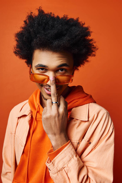 A young African American man with curly hair donning an orange shirt, jacket, and sunglasses against an orange backdrop, showcasing a vibrant and eclectic style. - Photo, Image