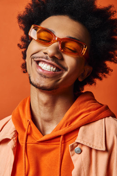 Стильный молодой афроамериканец с вьющимися волосами, модной одеждой и солнечными очками, улыбающийся на оранжевом фоне. - Фото, изображение