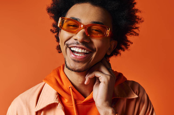 Ein junger, gelockter afroamerikanischer Mann in trendiger Kleidung und Sonnenbrille, der vor orangefarbenem Hintergrund ein strahlendes Lächeln in die Kamera wirft. - Foto, Bild