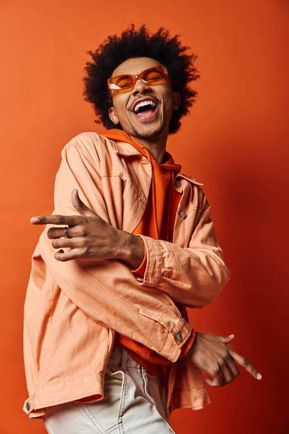 Homem afro-americano elegante com cabelo encaracolado usando óculos de sol, mostrando emoções em um fundo laranja. - Foto, Imagem