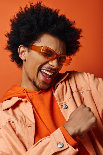 Κομψό Αφροαμερικανός άνδρας με αφρο μαλλιά σε πορτοκαλί πουκάμισο και γυαλιά ηλίου, αποπνέοντας δροσιά σε πορτοκαλί φόντο. - Φωτογραφία, εικόνα
