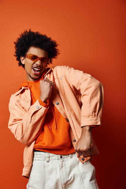 Κομψός Αφροαμερικανός με πορτοκαλί πουκάμισο και σακάκι, με γυαλιά ηλίου, σε πορτοκαλί φόντο. Μοντέρνο και εκφραστικό. - Φωτογραφία, εικόνα
