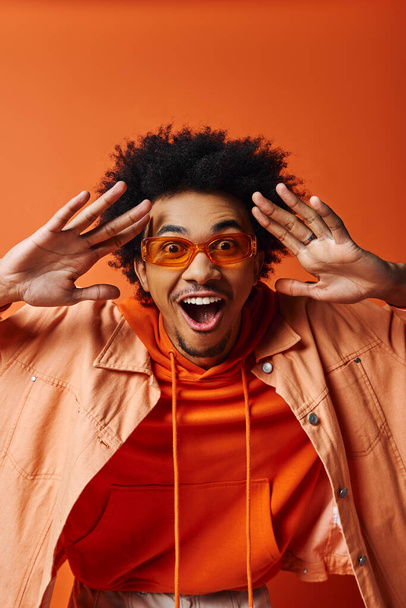 Stylish African American man with curly hair and trendy glasses making a funny expression on an orange background. - Photo, Image