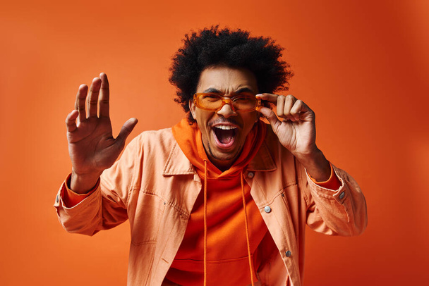 Tarz sahibi, kıvırcık saçlı ve güneş gözlüklü genç bir Afrikalı Amerikalı parlak bir arka planda turuncu gömlekle komik bir ifade sergiliyor.. - Fotoğraf, Görsel