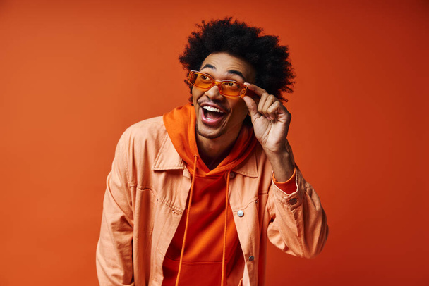 Een jonge Afro-Amerikaanse man met krullend haar in trendy kleding en zonnebril, met emoties tegen een oranje achtergrond. - Foto, afbeelding