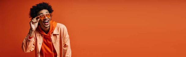 Un giovane afroamericano elegante, con i capelli ricci e l'abbigliamento alla moda, si copre gli occhi con una sciarpa arancione su uno sfondo arancione. - Foto, immagini
