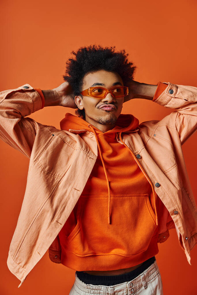 Stylový mladý Afroameričan s afro bundou a slunečními brýlemi, vyzařující chladnou a sebevědomou atmosféru na oranžovém pozadí. - Fotografie, Obrázek