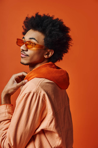 Un joven afroamericano elegante con el pelo rizado lleva gafas de sol y un atuendo de moda sobre un fondo naranja. - Foto, imagen