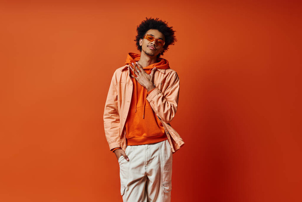 Un uomo afroamericano trendy e riccio posa in una giacca arancione e pantaloni bianchi su uno sfondo arancione audace, trasudando fiducia e stile. - Foto, immagini