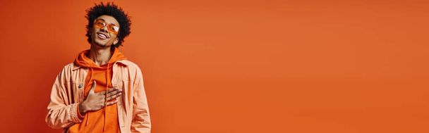 Un joven y elegante hombre afroamericano con el pelo rizado y gafas de sol con un traje naranja y blanco, exudando emoción. - Foto, Imagen