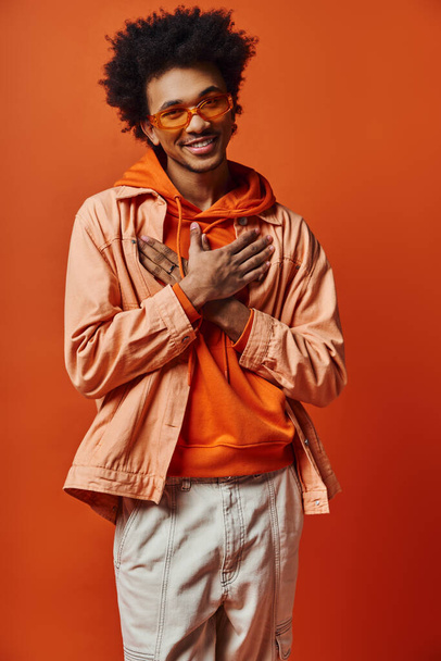 Kıvırcık saçlı, turuncu ceketli ve güneş gözlüklü genç bir Afrikalı Amerikalı parlak turuncu arka planda duruyor.. - Fotoğraf, Görsel
