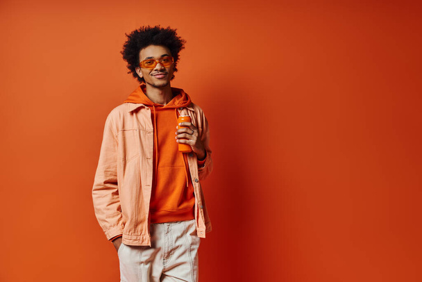 Un hombre afroamericano de moda en una sudadera con capucha naranja y gafas de sol disfrutando de una bebida sobre un fondo naranja vibrante. - Foto, imagen