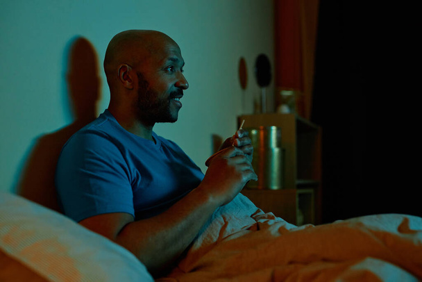 Portrait latéral d'un homme noir souriant regardant la télévision au lit la nuit, restant debout tard, copiant l'espace - Photo, image
