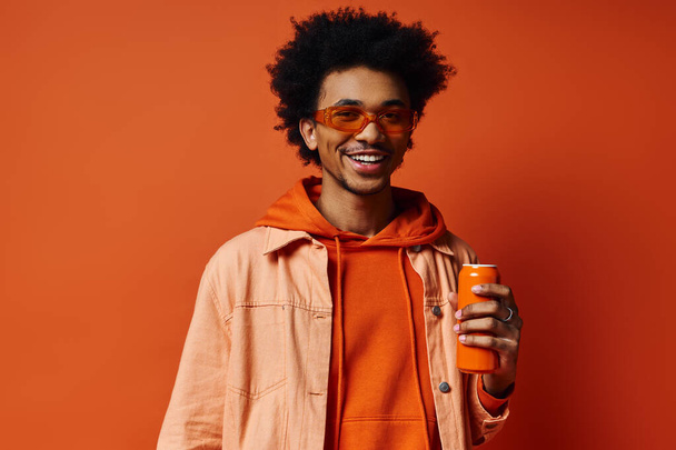 Trendy African American man in orange hoodie holding an orange juice can on vibrant orange background. - Photo, Image