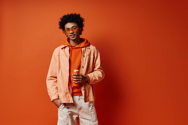 鮮やかなオレンジ色の背景にトレンディなオレンジのジャケットと白いズボンを自信を持ってポーズするスタイリッシュな若いアフリカ系アメリカ人男性. - 写真・画像